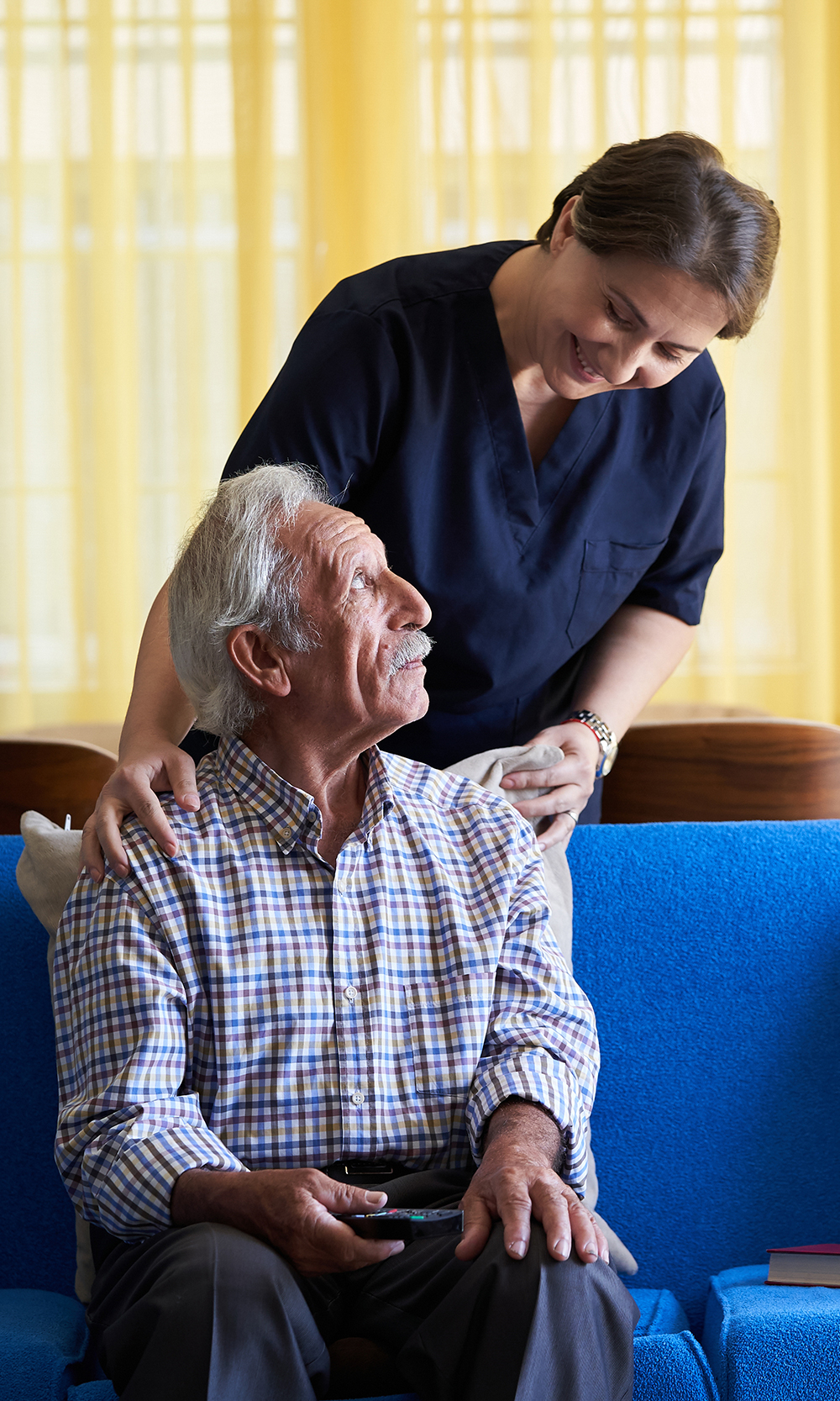 patient sitting on blue couch with caregiver standing checking on them